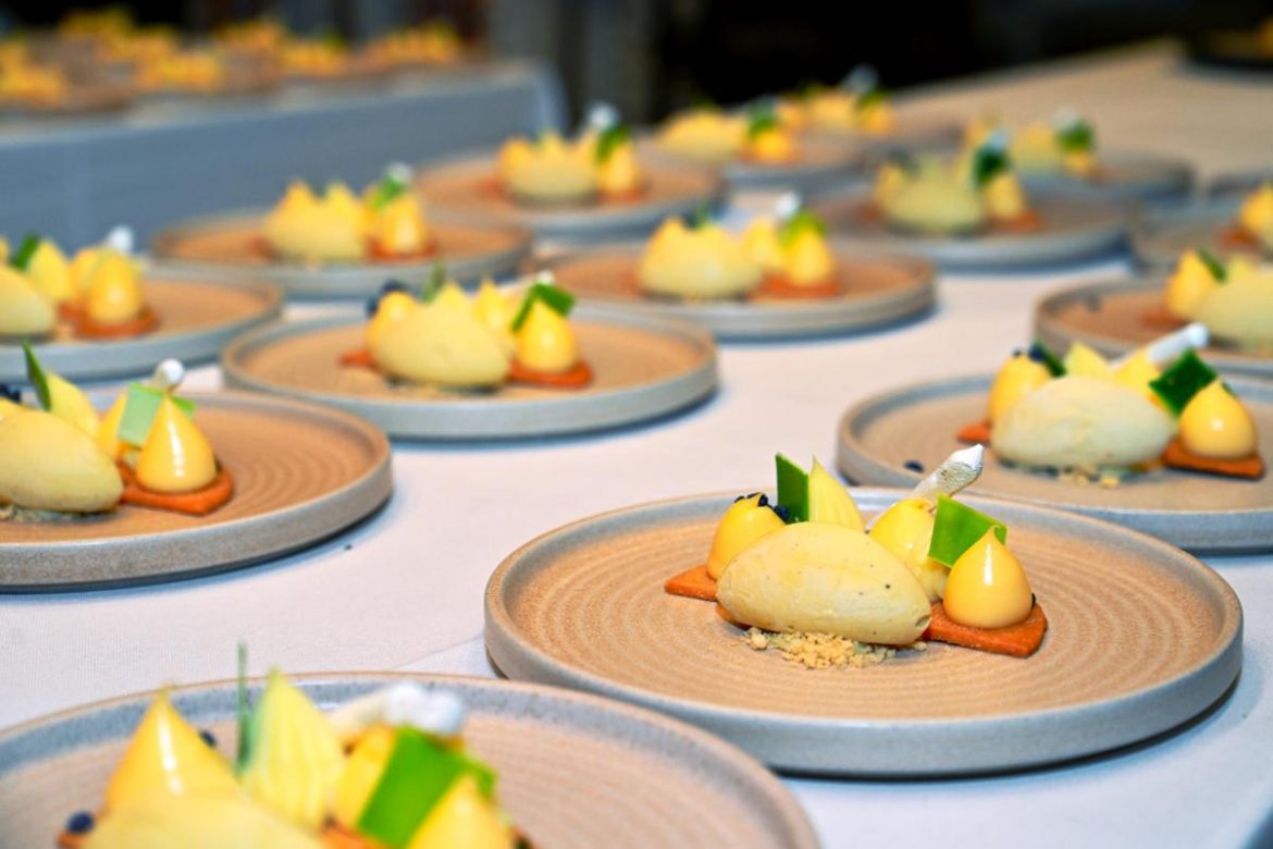 Catering multiple plates arranged on a table, each featuring a sophisticated dessert with ice cream kernel, small dollops of cream, green garnishes, and a rectangular biscuit base, presented on beige, textured ceramic plates.