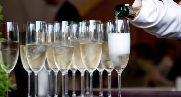 A row of elegant champagne flutes filled with chilled sparkling wine and ice, with a gloved hand pouring from a bottle into one of the glasses at a formal event catering.