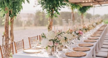 Romantic outdoor setup with a long table decorated with white catering tablecloths, pastel flower arrangements, crystal glasses, and gold-rimmed plates, under a pergola adorned with string lights and greenery.