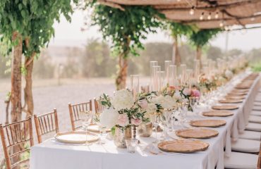 Romantic outdoor setup with a long table decorated with white catering tablecloths, pastel flower arrangements, crystal glasses, and gold-rimmed plates, under a pergola adorned with string lights and greenery.