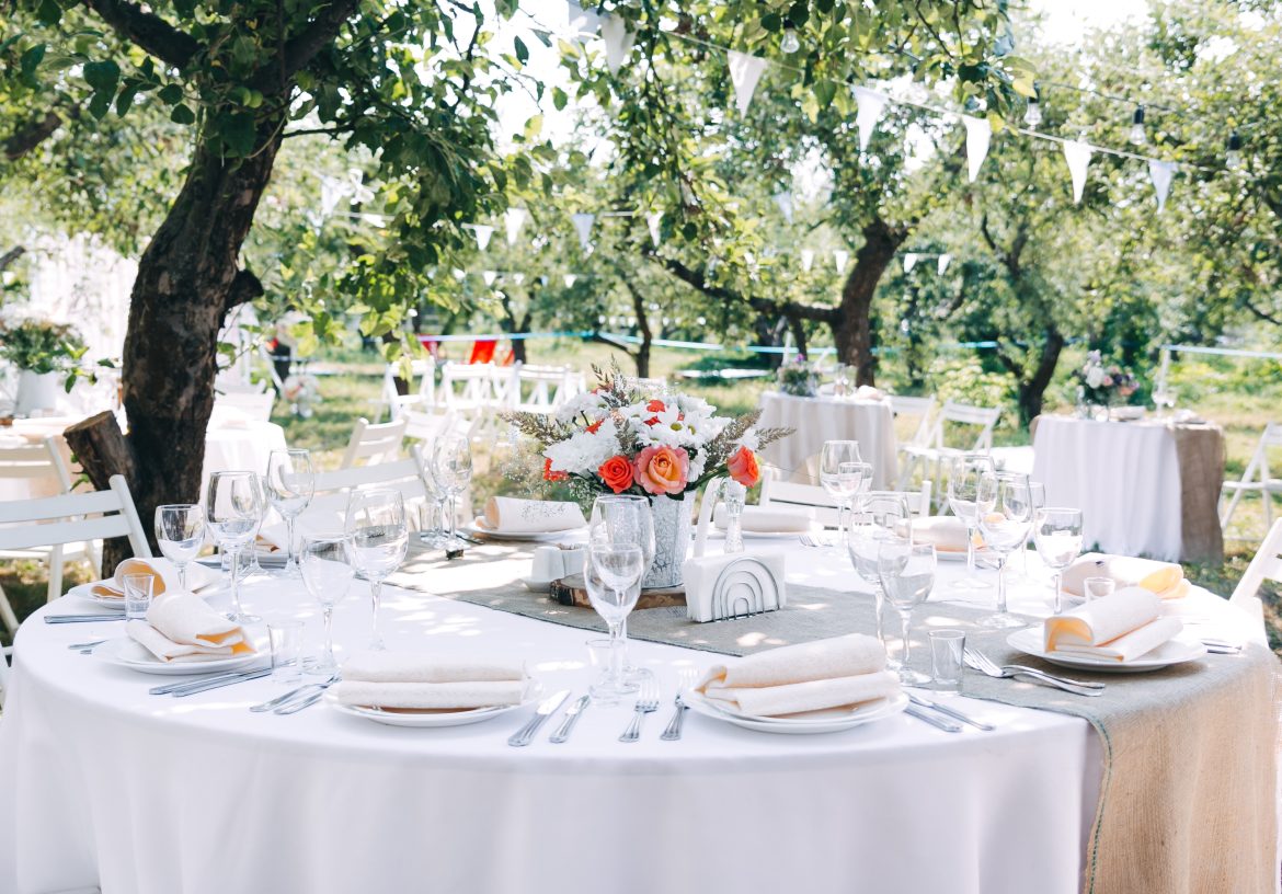 Outdoor reception with round tables, white tablecloths, floral centerpieces, and elegant decoration under trees – event catering in a natural setting.