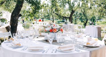 Outdoor reception with round tables, white tablecloths, floral centerpieces, and elegant decoration under trees – event catering in a natural setting.