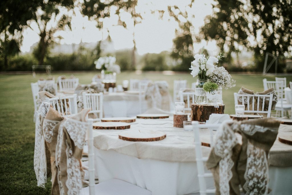 Outdoor reception with round tables, white tablecloths, floral centerpieces, and elegant decoration under trees – event catering in a natural setting.