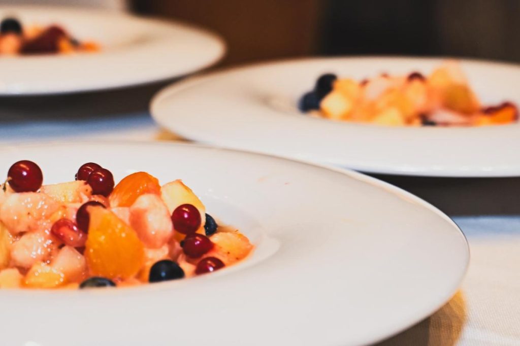 Catering multiple plates arranged on a table, each featuring a sophisticated dessert with ice cream kernel, small dollops of cream, green garnishes, and a rectangular biscuit base, presented on beige, textured ceramic plates.