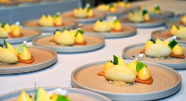 Catering multiple plates arranged on a table, each featuring a sophisticated dessert with ice cream kernel, catering, small dollops of cream, green garnishes, and a rectangular biscuit base, presented on beige, textured ceramic plates.