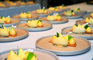 Catering multiple plates arranged on a table, each featuring a sophisticated dessert with ice cream kernel, catering, small dollops of cream, green garnishes, and a rectangular biscuit base, presented on beige, textured ceramic plates.