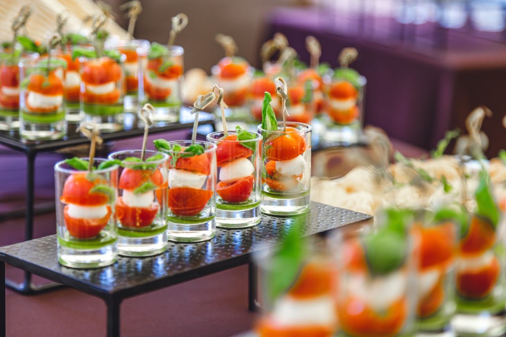 Ποικιλία από καλοστημένα finger food σε ποτήρια και πιατάκια, με υλικά όπως σολομός, μοτσαρέλα, ντοματίνια και φρέσκα μυρωδικά, σε τραπέζι catering.