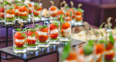 A variety of elegantly presented finger foods in glasses and small plates, featuring ingredients like salmon, mozzarella, cherry tomatoes, and fresh herbs, on a catering table.