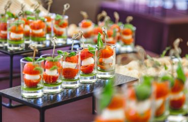 A variety of elegantly presented finger foods in glasses and small plates, featuring ingredients like salmon, mozzarella, cherry tomatoes, and fresh herbs, on a catering table.