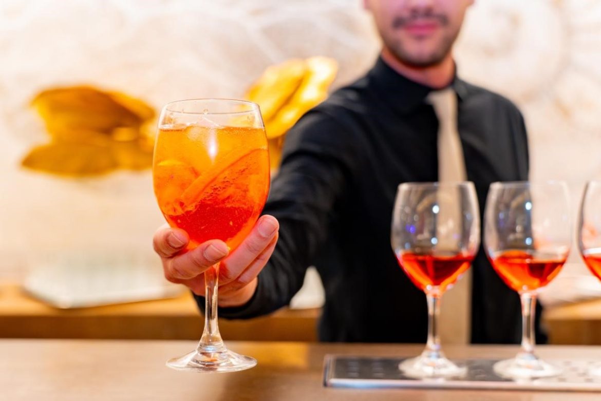 A bartender catering in a black shirt and beige tie serves an orange cocktail with ice and a slice of orange, with more filled glasses lined up on the bar.
