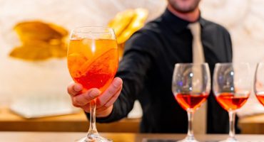 A bartender catering in a black shirt and beige tie serves an orange cocktail with ice and a slice of orange, with more filled glasses lined up on the bar.