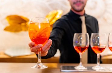 A bartender catering in a black shirt and beige tie serves an orange cocktail with ice and a slice of orange, with more filled glasses lined up on the bar.