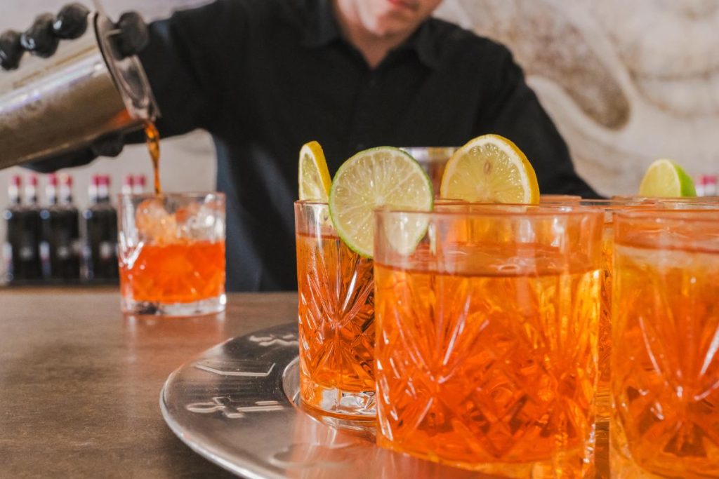 A bartender catering in a black shirt and beige tie serves an orange cocktail with ice and a slice of orange, with more filled glasses lined up on the bar.