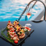 Πιατέλα με καλοκαιρινά finger foods δίπλα σε πισίνα, με ποτήρι αφρώδους οίνου, ιδανικό σκηνικό για pool party catering.